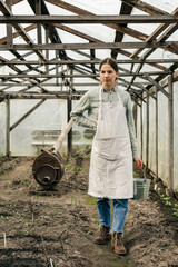 An attractive woman holding a tin bucket and walking in the greenhouse looking at seedlings