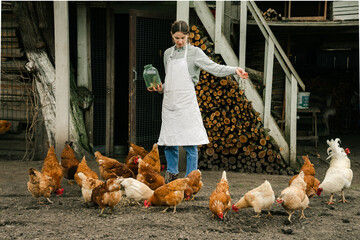 an attractive young woman standing near the chicken coop holding a jar with grain and feeding the...