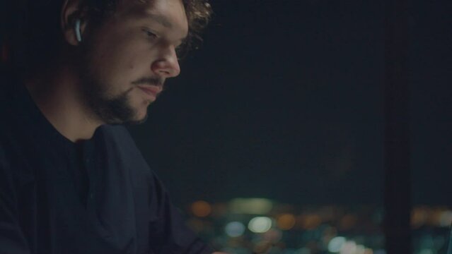 A curly-haired white man in a black T-shirt and a watch typing text illuminated screen late at night against bokeh lights of a night city.