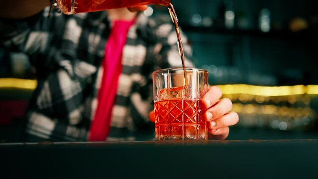 Experienced bartender pouring negroni alcoholic cocktail with crystal ice cubes from measuring glass into glass at bar