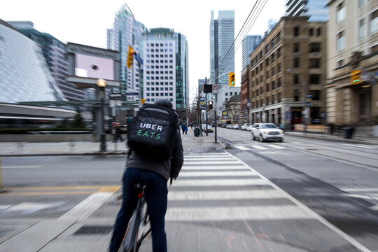 TORONTO, CANADA - DECEMBER 31, 2016: Uber Eats delivery man on a bicycle waiting to cross a street in the center of Toronto, Ontario, with motion blurs. Uber Eats is one of the leader of the online fo