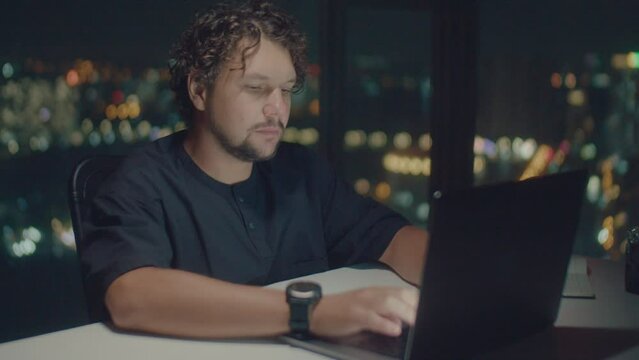 Сlose-up shot of a curly-haired European man wearing black T-shirt and sport watch works at a computer on the background of a panoramic window with a view of lights of the city in the bokeh