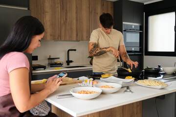 Two young latin people using the smartphone while cooking traditional arepas and llapingachos.