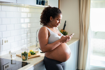 Young pregnant latina woman using a smart phone in the kitchen of a home