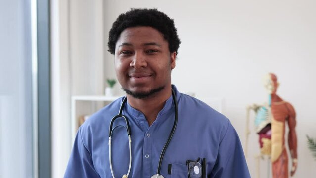 Portrait of attractive multicultural man in blue scrubs and stethoscope standing in doctor's office with human anatomy model on background. Cheerful surgeon posing in medical center interior.