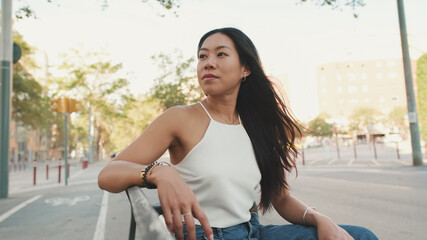 Pretty brunette girl, dressed in white top and jeans, sits on park bench, enjoys the good weather, free time