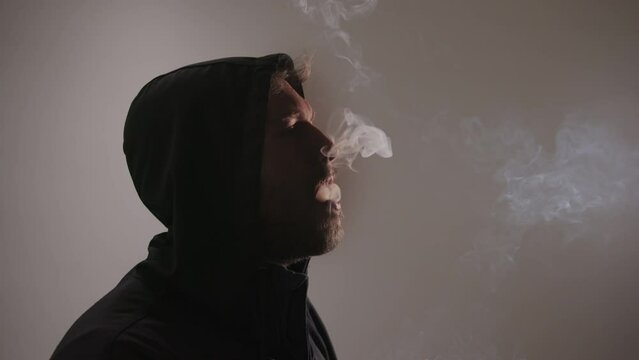 A man in a hoodie enjoying a cannabis joint: closeup of his face as he inhales and exhales the smoke. Marijuana culture and lifestyle.
