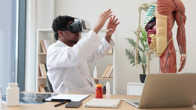 African american man wearing lab coat and 3d goggles using VR system while sitting near modern devices and human skeleton in office. Therapist preparing for medical training in teaching hospital.