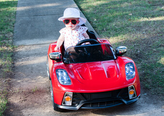 Caucasian baby in a white hat riding in an electric convertible red sport car. child driving a car on a summer day