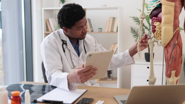 Confident multicultural adult in lab coat pointing at femur of human anatomy model while holding tablet in doctor's office interior. Physician collecting information for patient education.
