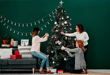That should go there. three attractive middle aged women decorating a Christmas tree together at home.