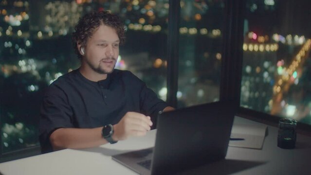 A curly-haired white man in a black T-shirt and a watch explains how to trade on the stock exchange late at night against the background of a glass panoramic corner and a night city.