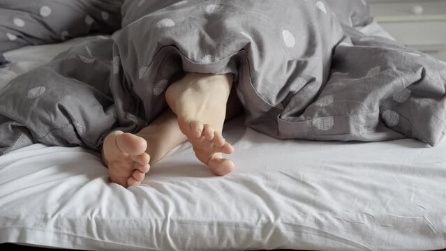 Closeup of female feet lying on bed and showing from under the blanket, woman turning around and searching for comfortable pose. Concept of relaxing at home, comfortable bed, resting in morning