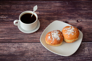 Two fresh donuts with powdered sugar and a cup of hot black coffee on a wooden table.