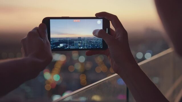 Close-up of woman's hands holding smartphone and taking pictures or video of amazing orange city sunset. Beautiful panoramic view of sundown