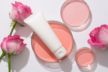 Obraz na płótnie Canvas Scene for branding cosmetic of rose extract, plastic tube unlabeled for design placed on petri dish containing pink liquid and fresh roses arranged on white background