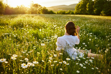 a red-haired woman in a light dress sits in a chamomile field at sunset and admires the passing day