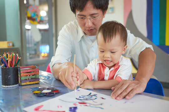 Cute smiling little Asian toddler baby boy child painting with brush and watercolors, Businessman father painting with son after working time, Creative play for toddlers, Soft and selective focus