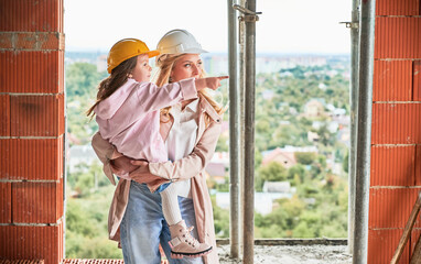 Woman in building helmet holding child while standing at construction site. Kid pointing away. Mother and daughter posing in apartment building under construction. - obrazy, fototapety, plakaty