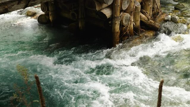 The powerful stream of a clear mountain river and the support of a wooden bridge made of logs. The water in river is turquoise in colour. Camera movement from bottom to top. Slow motion