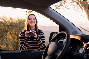young caucasian woman laughing happy with a photo camera in hands during a road trip in camper van, concept of van life and travel adventures