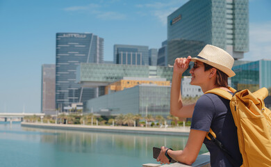 Fototapeta na wymiar travel to the United Arab Emirates, Happy young asian female traveler in dress and hat look at View of Abu Dhabi Skyline in Al Maryah Island. Vacation and tourist destination concept.