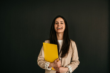 Portrait of a smiling businesswoman holding files and standing against the black background. Studio...