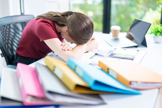 Very stressed business woman and female employee sitting in front of her computer with her hands in front of her eyes, feeling sad and depressed. Too much work - huge pile of paperwork at office table