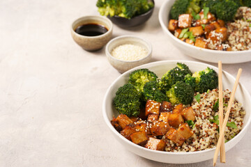 Two quinoa bowls with steamed broccoli and smoked tofu cubes in teriyaki sauce with sesame seeds as topping on white checkered kitchen napkin, grey concrete background