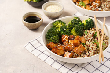 One quinoa bowl with steamed broccoli and smoked tofu cubes in teriyaki sauce with sesame seeds as topping on white checkered kitchen napkin, grey concrete background