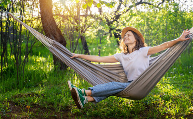 Young happy caucasian woman in a hat lying in a hammock in a green garden enjoying a summer day