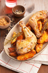 two oven-roasted organic chicken thighs on roasted potato wedges spiced with chili and paprika on oval ceramic plate, rosemary and black pepper. Checkered kitchen towel, brown wooden tabletop