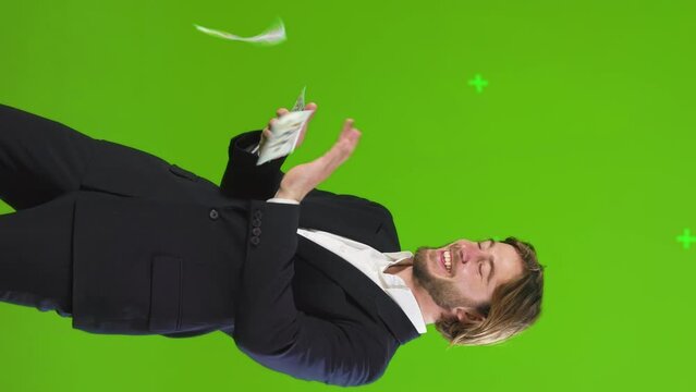 Wealthy cheerful businessman throwing cash in air, having fun in studio. Side view of confident rich man wearing suit, scattering money around, smiling, motion, isolated on green. Lifestyle concept.