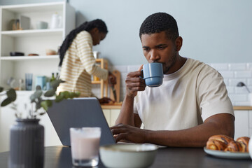 African American young man using laptop during breakfast at table in the kitchen with his girlfriend cooking in background