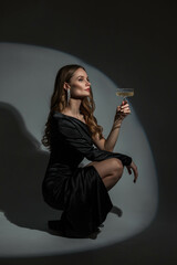 Elegant beautiful vogue chic woman model in black dress with vintage glass of wine sits and poses...