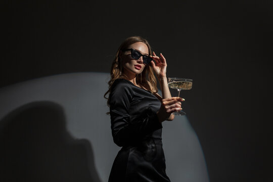 Fashionable Beautiful Elegant Vogue Woman Wears Cool Sunglasses In A Fashion Evening Black Dress With A Glass Of Champagne At A Party. Chic Pretty Lady Celebrates An Event