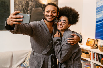 Fototapeta na wymiar Cheerful couple taking selfie and hugging while standing in living room