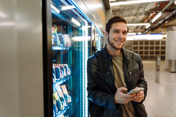 Smiling man using mobile phone while standing near snacks vending machine in subway - obrazy, fototapety, plakaty