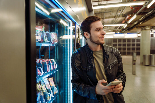 Smiling man using mobile phone while standing near snacks vending machine in subway