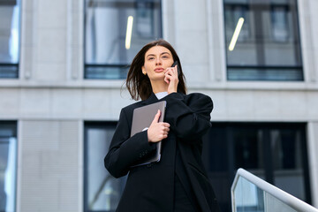 Fototapeta premium Portrait of a businesslike young woman who is talking on the phone and holding a laptop in her hands against the background of the office