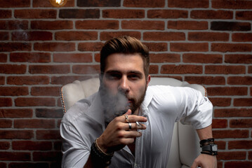 A stylish Arab guy is having a business meeting while sitting in a cozy chair near a red brick wall. An oriental guy is very focused, with a cigarette in his mouth and smoking