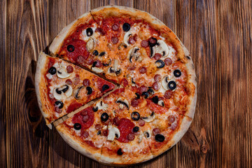 Tomato-based pizza with mozzarella cheese, hunting sausages, salami, olives, mushroom sauce. On a wooden background