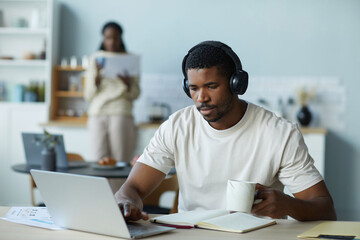 African American man in wireless headphones drinking coffee and working online on laptop at home