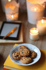 Cup of tea or coffee, plate of cookies, books, e-reader, pencil and lit candles on the table. Selective focus.