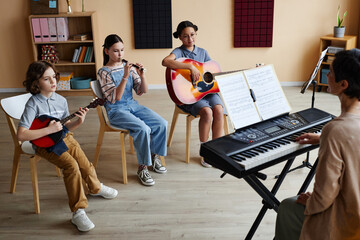 Group of children sitting on chairs and playing musical instruments with teacher playing piano during lesson in music class - Powered by Adobe