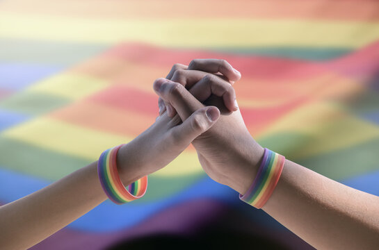 Closeup view of hands of gay couple wearing rainbow wristbands to present LGBT love, blurred rainbow background, concept for LGBT people celebrations in pride month.