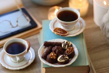 Books, reading glasses, e-reader, plate of chocolate pralines, bowl of cookies, cups of tea and lit candles on the table. Selective focus.