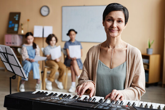 Portrait of mature music teacher smiling at camera while playing piano in music class with children in background
