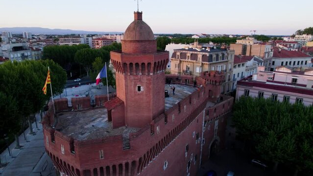 Drone clip encircling Le Castillet Fortress in Perpignan, France overhead at sunset, with musician playing on rooftop and French flag flying in gentle breeze