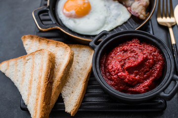 Fried eggs on a stylish ceramic plate with handles with fried bacon, cherry tomatoes, tomato paste and bread toast on a dark background. Breakfast, lunch and dinner. Delicious food.